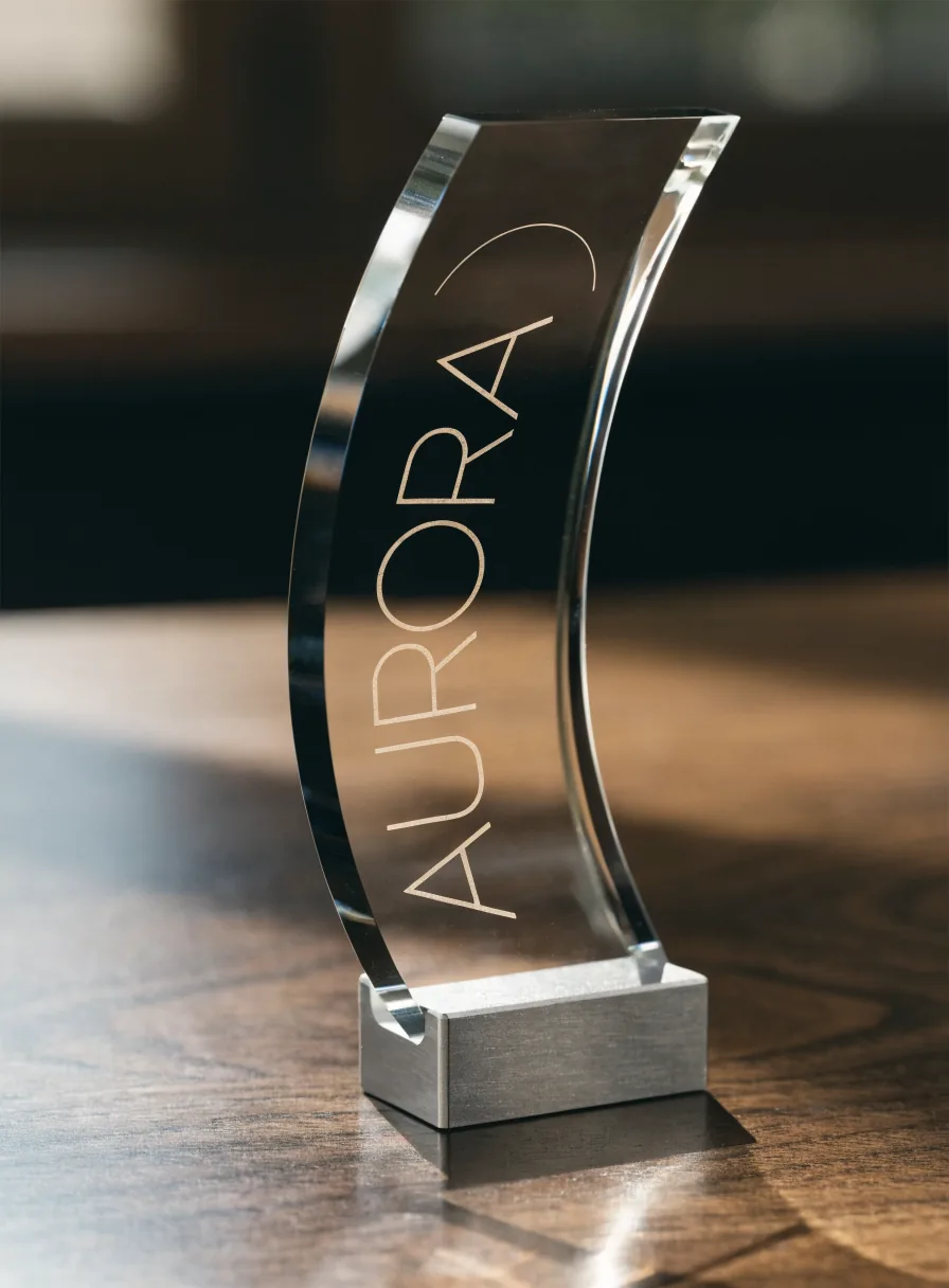 placa de cristal óptico grueso curvada para sostenerse sola, montada en una zapata de aluminio maquinado sobre un escritorio de nogal, con la palabra Aurora grabada a láser en línea fina sobre la cara y un arco de amanecer grabado arriba