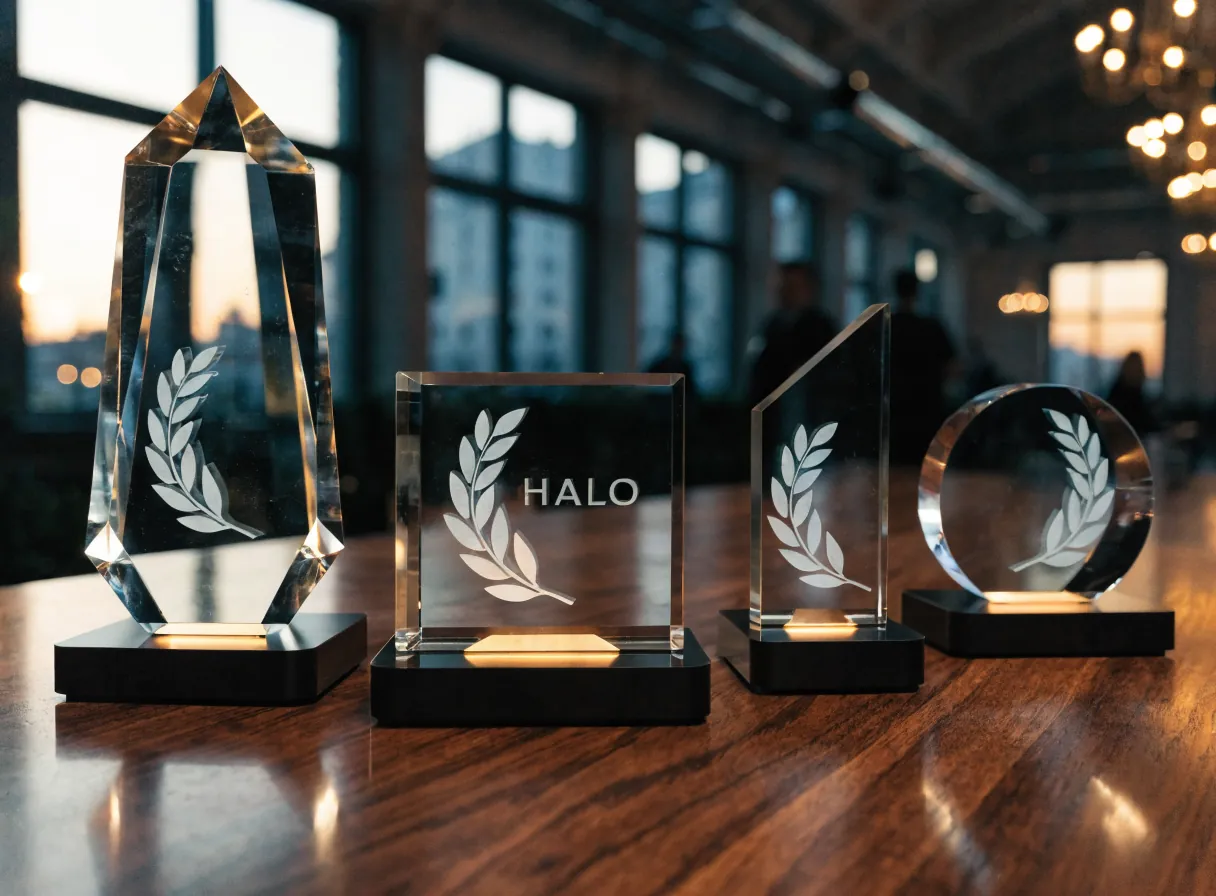 cuatro trofeos de cristal de alturas distintas sobre una mesa de nogal, cada uno en una base negra encendida que sube la luz por el cristal, el del centro grabado con la palabra Halo