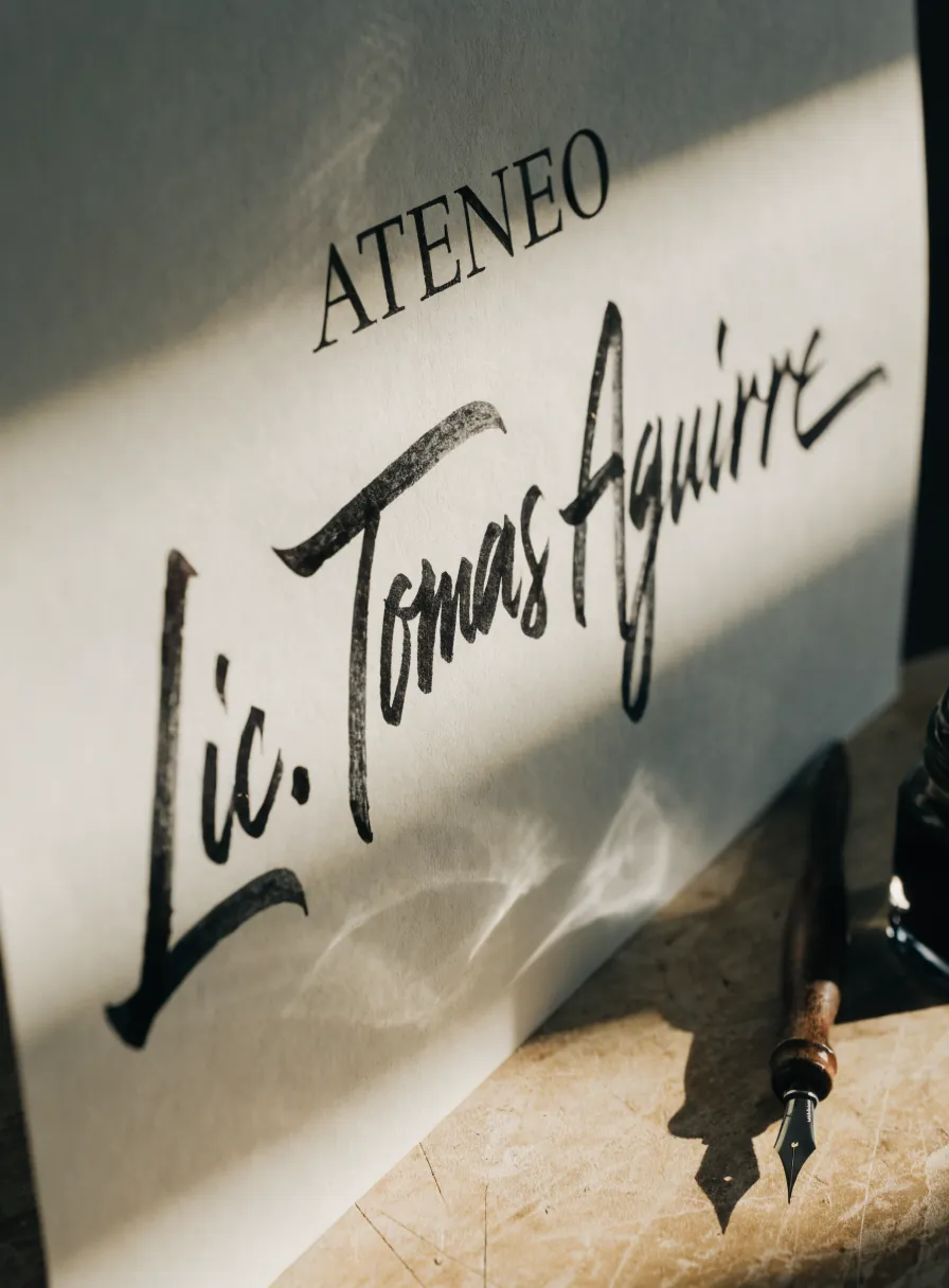 nombre Lic. Tomás Aguirre trazado a mano con plumilla sobre un diploma de algodón, con la pluma de madera apoyada junto a la hoja