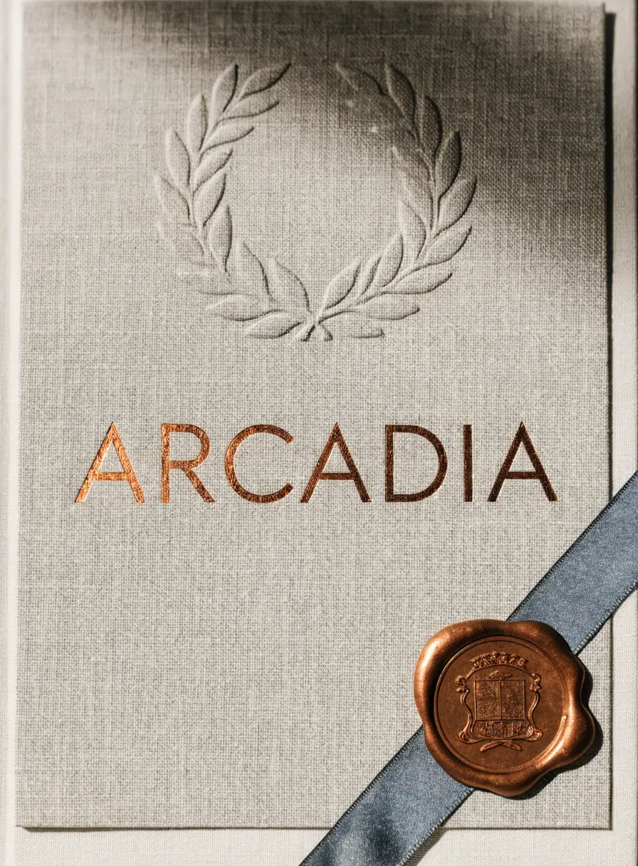 diploma en papel de lino gris humo visto desde arriba, con la palabra Arcadia estampada en foil cobre, una corona de laurel prensada en realce ciego profundo arriba y un sello de cera cobre prensado sobre un listón de seda gris en la esquina