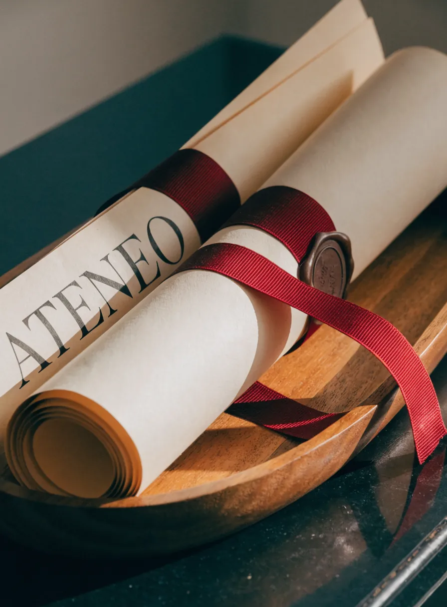 dos diplomas enrollados en una charola de madera, uno atado con listón rojo y sello de cera y el otro mostrando la palabra Ateneo