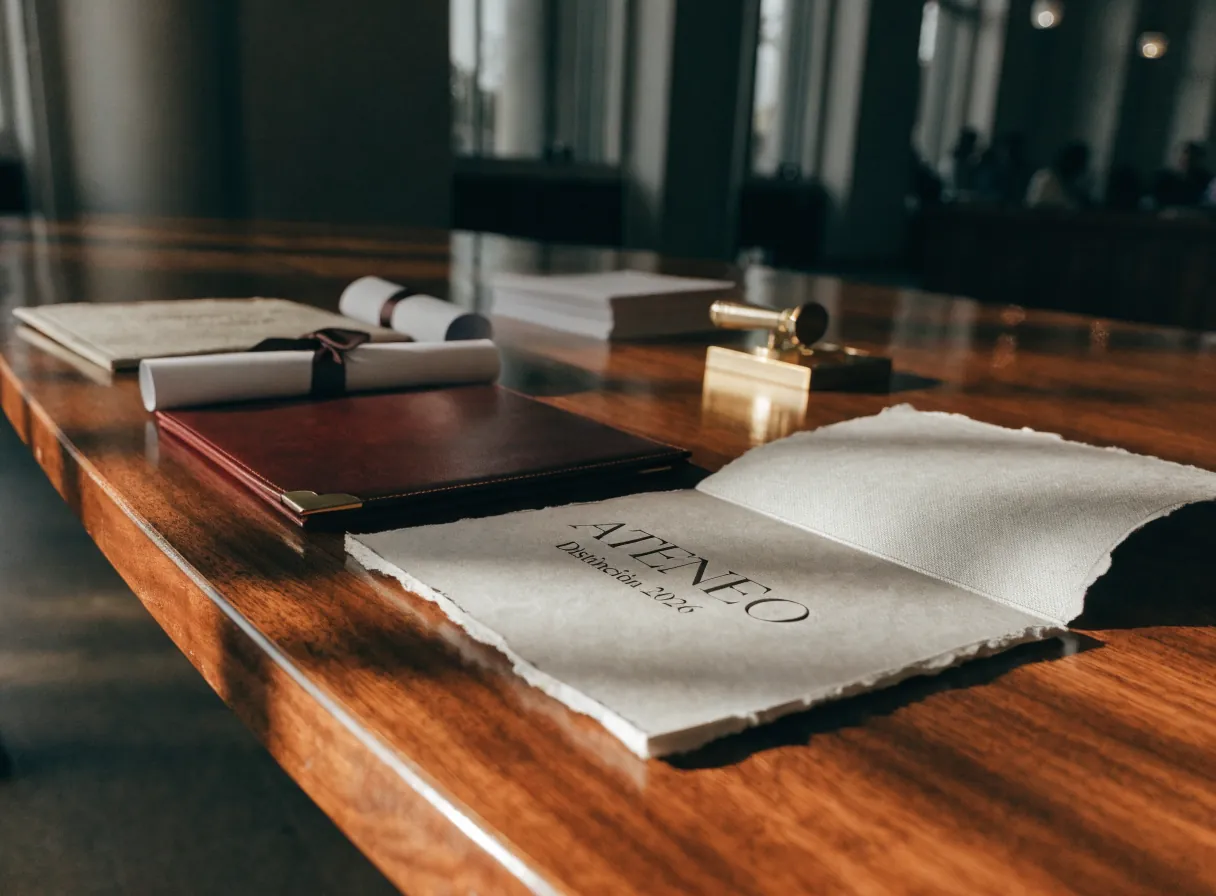 mesa de ceremonia con cuatro diplomas, uno abierto en papel de algodón de borde irregular impreso con la palabra Ateneo, una carpeta de piel vino, dos diplomas enrollados y una prensa de sello dorada