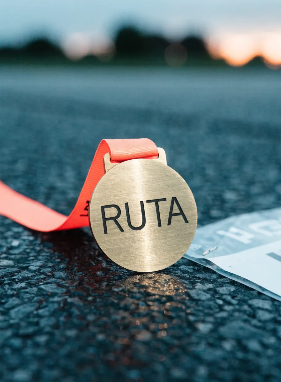 medalla redonda de latón con la cara cepillada y la palabra RUTA acuñada, sobre el asfalto mojado al amanecer junto a un número de corredor doblado, con cinta impresa color coral