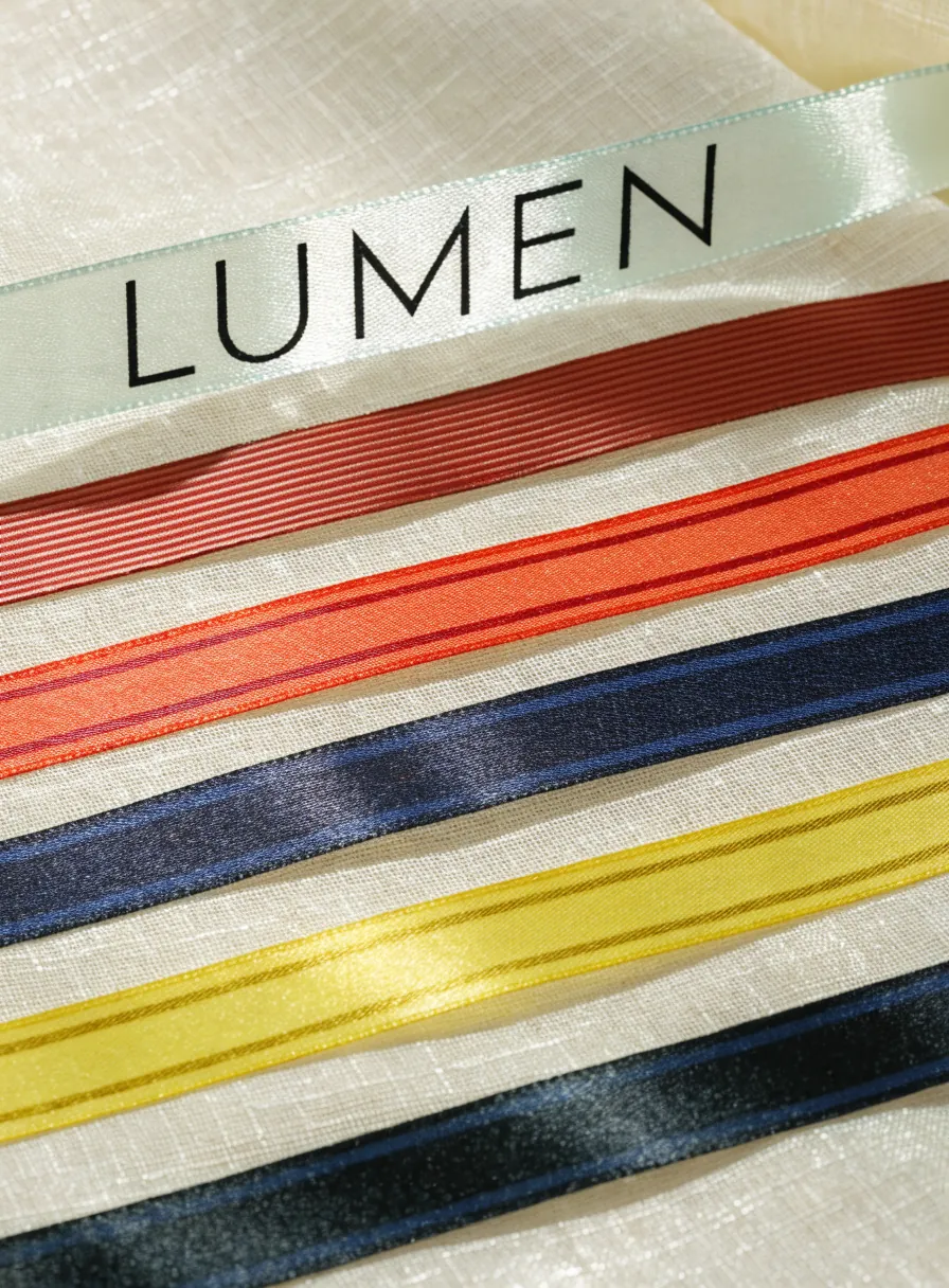 cinco cintas planas para medalla en colores y patrones distintos, extendidas y encimadas sobre una superficie de lino claro, con la palabra LUMEN impresa a lo largo de la primera