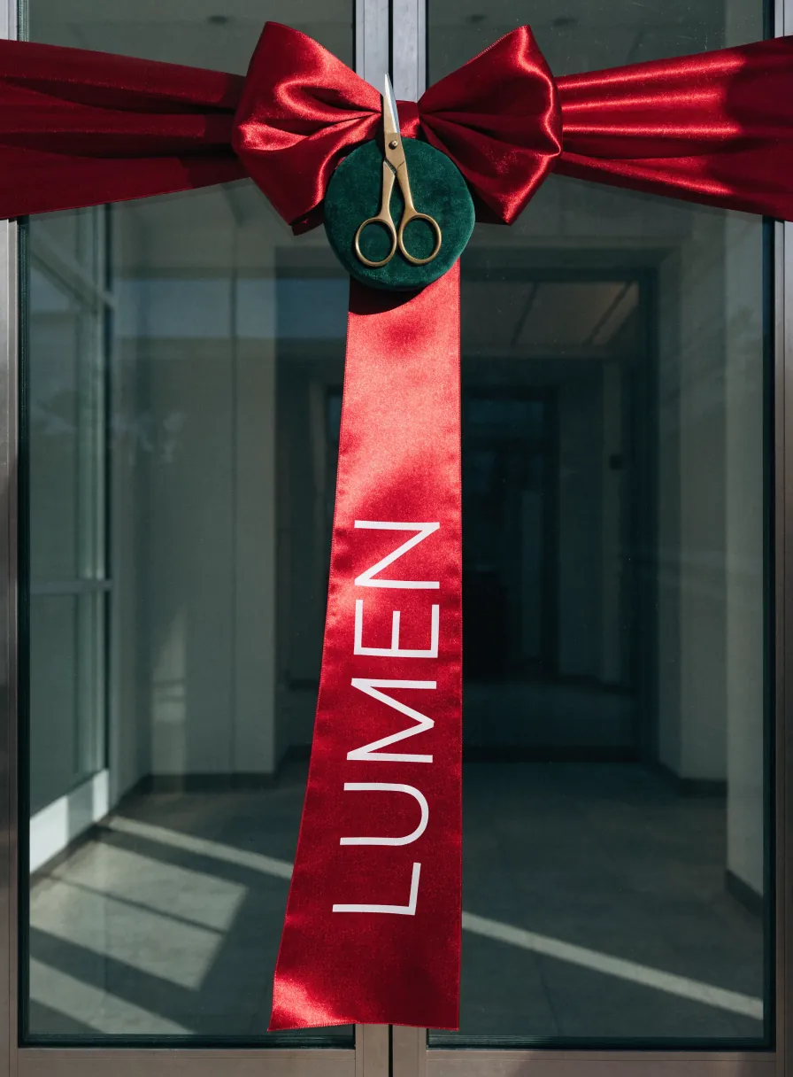 listón ancho de satín rojo atado en moño sobre las puertas de cristal de un edificio nuevo, con unas tijeras doradas de ceremonia colgando de un disco de terciopelo verde y la palabra LUMEN impresa en blanco a lo largo de la cola del listón