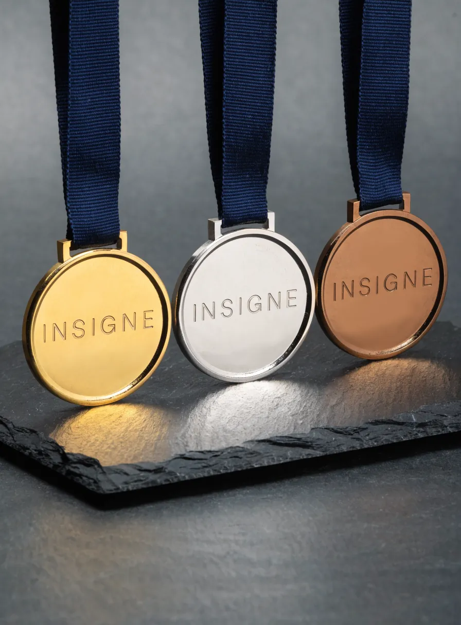 tres medallas del mismo diseño con baño de oro, plata y bronce, alineadas sobre una losa de pizarra oscura, cada una con su cinta tejida azul marino y la palabra INSIGNE acuñada