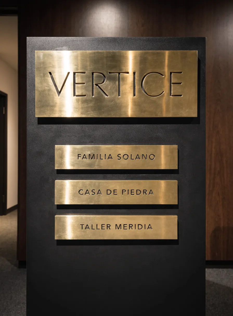 panel de metal negro mate en el vestíbulo de una fundación, con una placa cabecera de latón grabada VERTICE y tres placas nominales grabadas FAMILIA SOLANO, CASA DE PIEDRA y TALLER MERIDIA